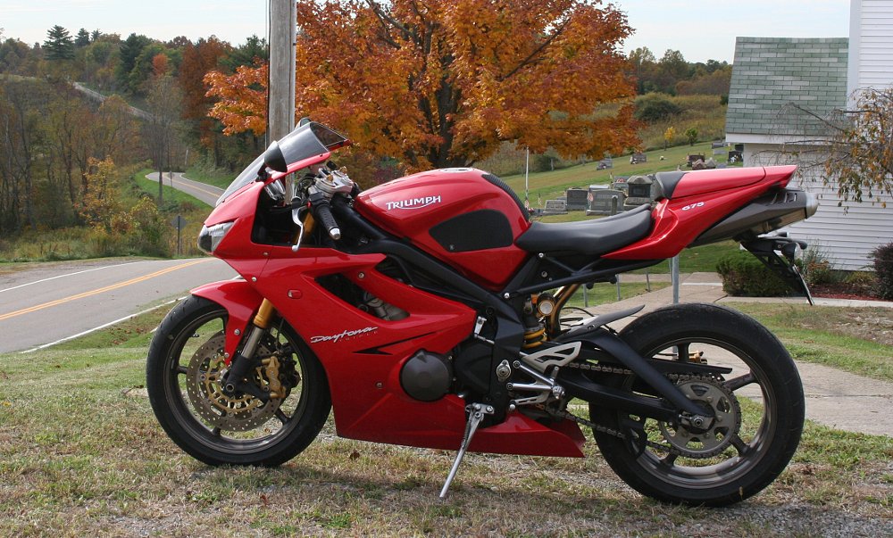 Red Triumph Daytona parked by a white country church in the fall with a road snaking off over the hills