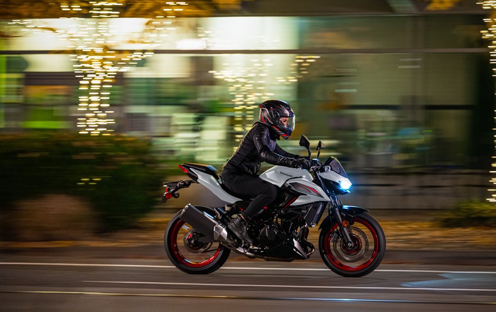woman rider on a white Z500 on a city street at night, riding past a storefront of bright lights