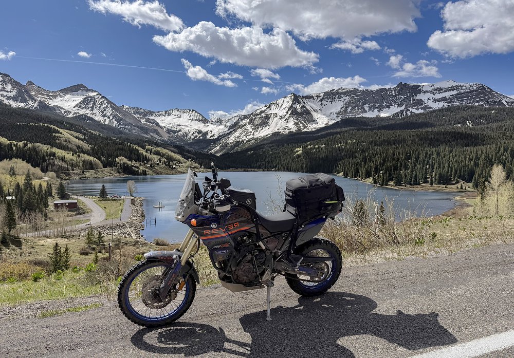 motorcycle parked at an overlook above a mountain lake with snowy mountains in the distance under a bright blue sky