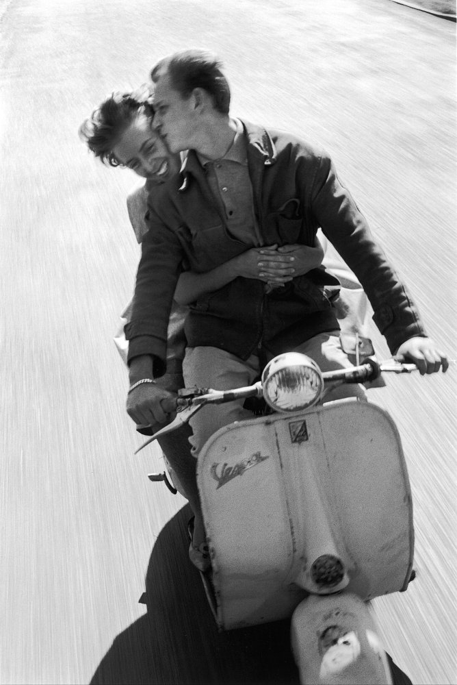 old black-and-white photo of a man and woman riding a Vespa, the man kissing the woman on the cheek