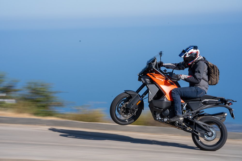 2027 KTM 1390 Super Adventure S EVO wheelying on a mountain road with a blurred background.