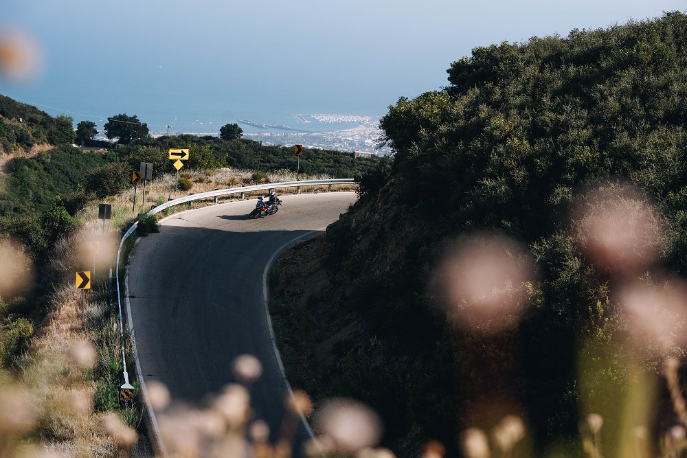 2027 KTM 1390 Super Adventure S EVO riding through a curve on a twisty mountain road with the ocean and Santa Barbara, California in the background.