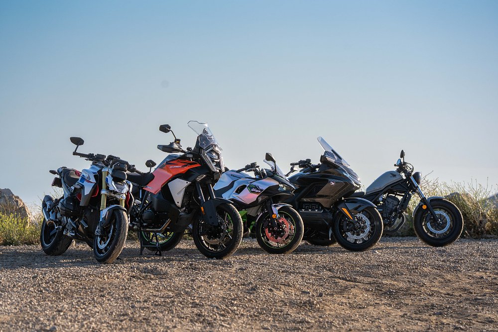 BMW's R 1300 R, KTM's 1390 Super Adventure S EVO, Kawasaki's Ninja 7 Hybrid, and Honda's Gold Wing Tour parked next to each other in a gravel lot..