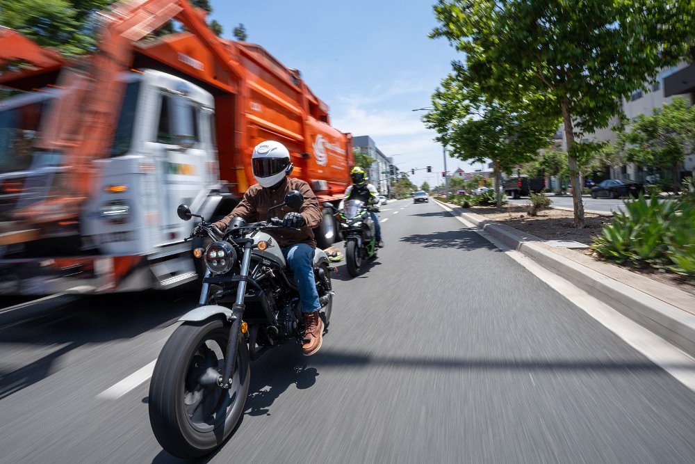 A Honda Rebel 300 and Kawasaki Ninja 7 Hybrid ride past a garbage truck on a city street.