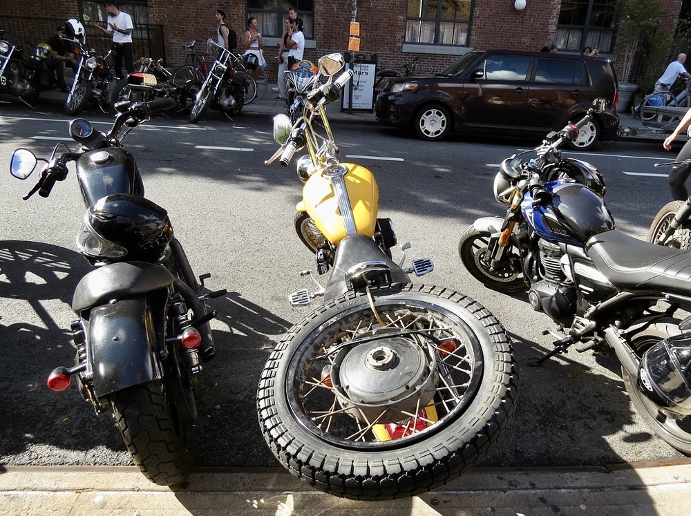 a vintage yellow motorcycle with a spare wheel mounted on the back is among the motorcycles parked along the street outside the show