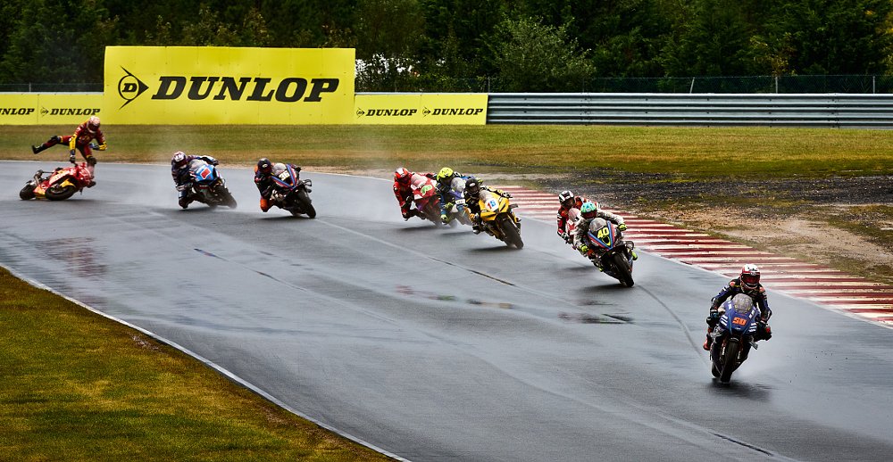 Bobby Fong on his blue Yamaha leads a field of races through a series of curves and at the back of the field Tyler Scott is thrown in the air by a highside crash