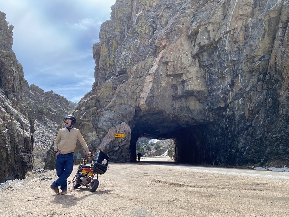 A man and a mini bike on the side of a twisty road with the road running through a rock tunnel in the background