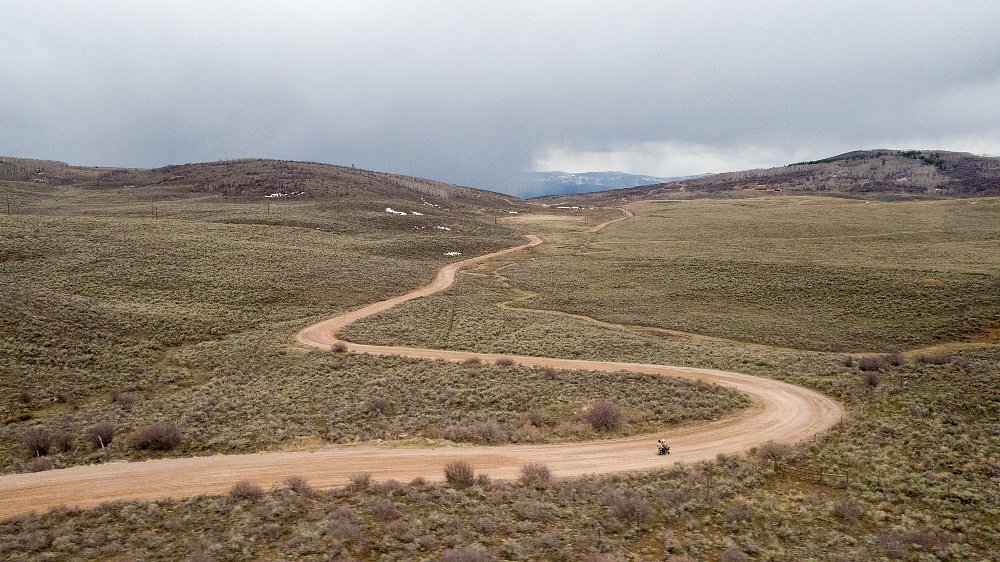 A gravel road snakes through low hills with rain on the horizon, and a mini bike riding toward it.