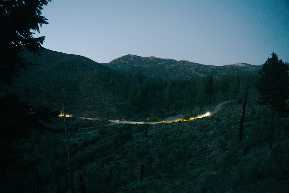 A far away shot shows a scene at dusk in the pine trees with headlights illuminating the trail as night riders ride on the trail.