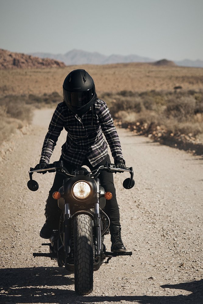 A rider is an in awkward standing position on the footpegs of the Indian Scout Bobber on a rocky dirt road.