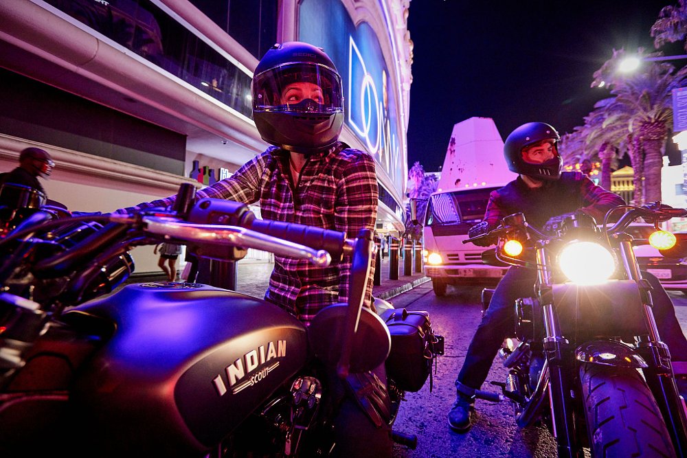 A group of cruiser bikes and riders sit on the Las Vegas Strip at nightime with bright purple lights.