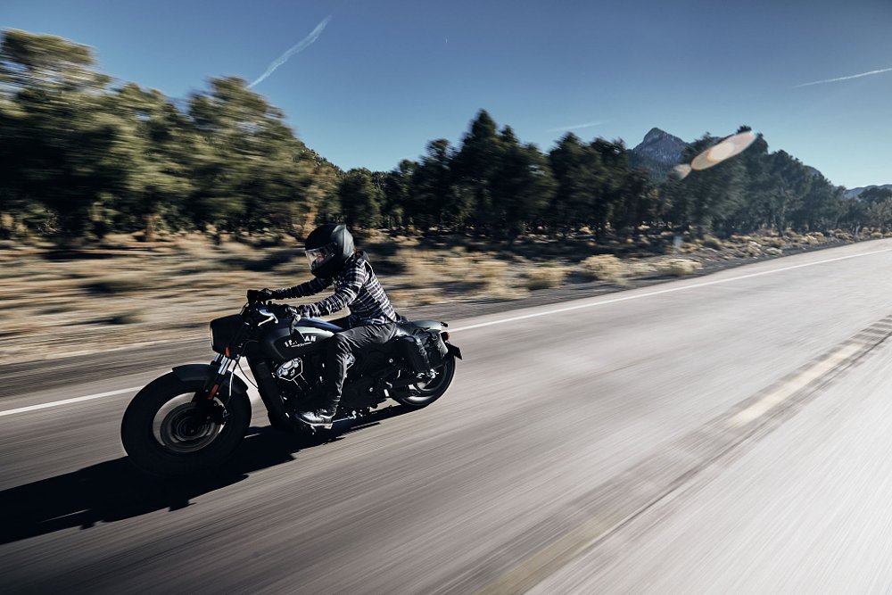 A female rider rides the Indian Scout Bobber motorcycle down a forest road.