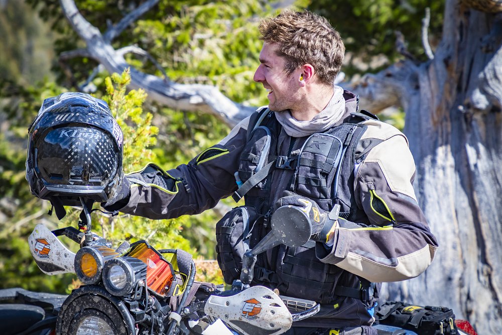 A rider stand by his bike wearing the Klim pack vest on a wooded trail.