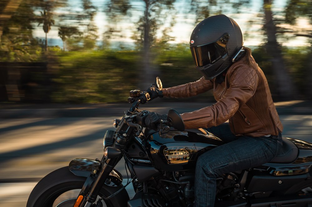 A waist up close up of a female rider in brown leather jacket, black helmet, hair in the wind on board the Sportster S