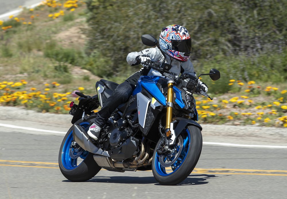 A front 3/4 view of a female rider piloting the GSX-S1000 on a winding back country road with flowers