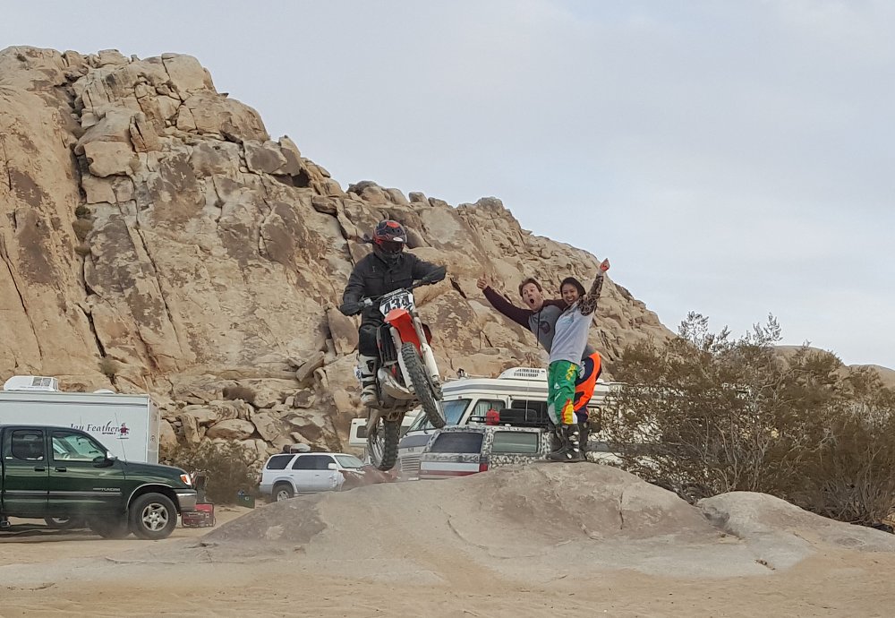 A dirt bike rider hits a small jump while friends cheer him on