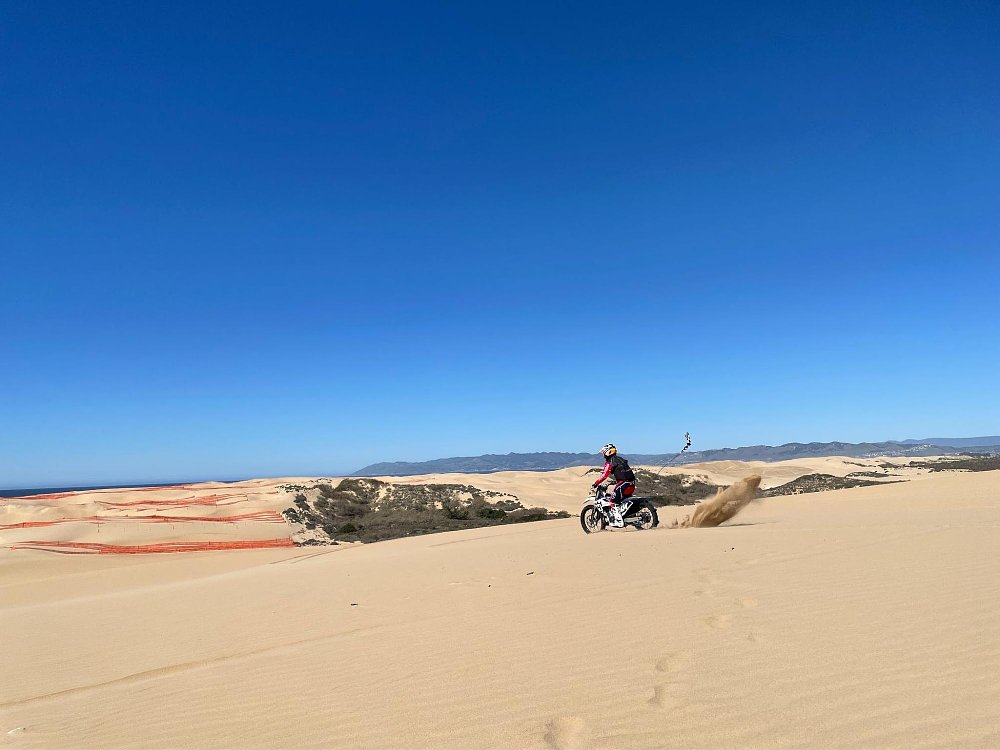 As a dirt rider rides in the dune sand, you can see bright orange plastic fencing in the background meant to mitigate dust