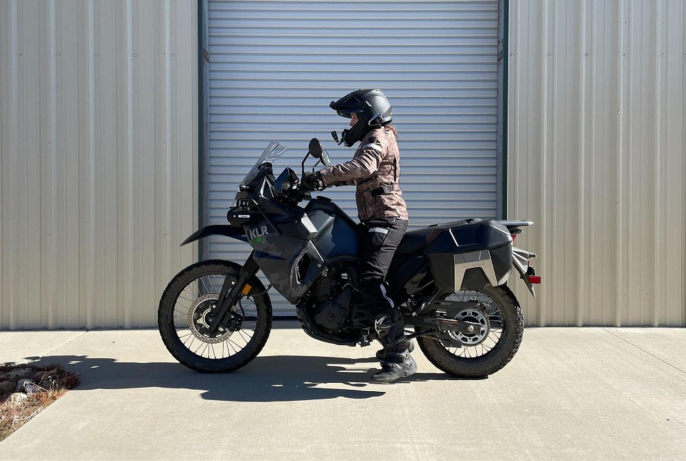 A short female rider with legs dangling while sitting on the full size KLR650 motorcycle