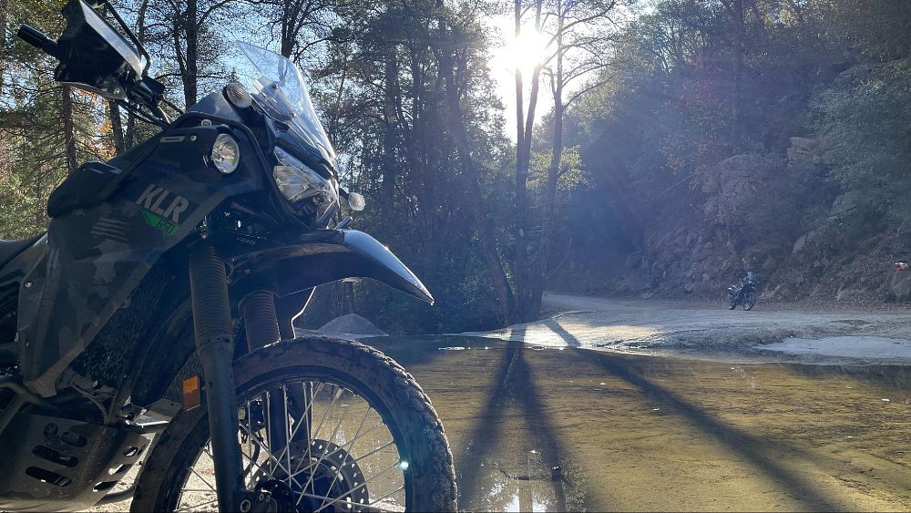 The KLR650 Adventure sits in foreground, with the KLR650 S in the background in a pond reflection