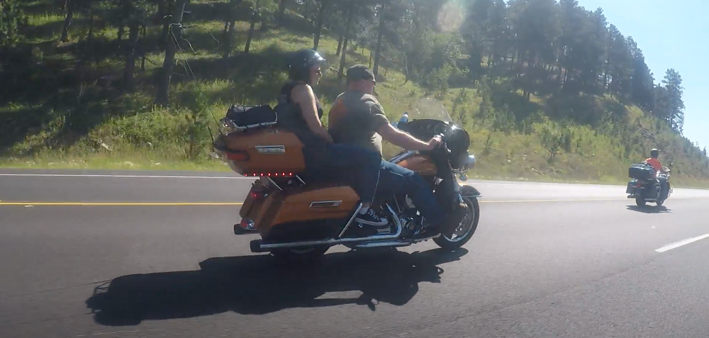 a motorcycle rider in sturgis rides with no helmet on, while his passenger does wear a helmet