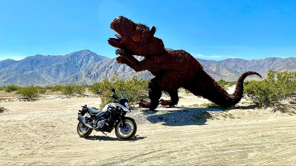 A farway view of the V-strom parked next to metal dinosaur sculpture in desert landscape