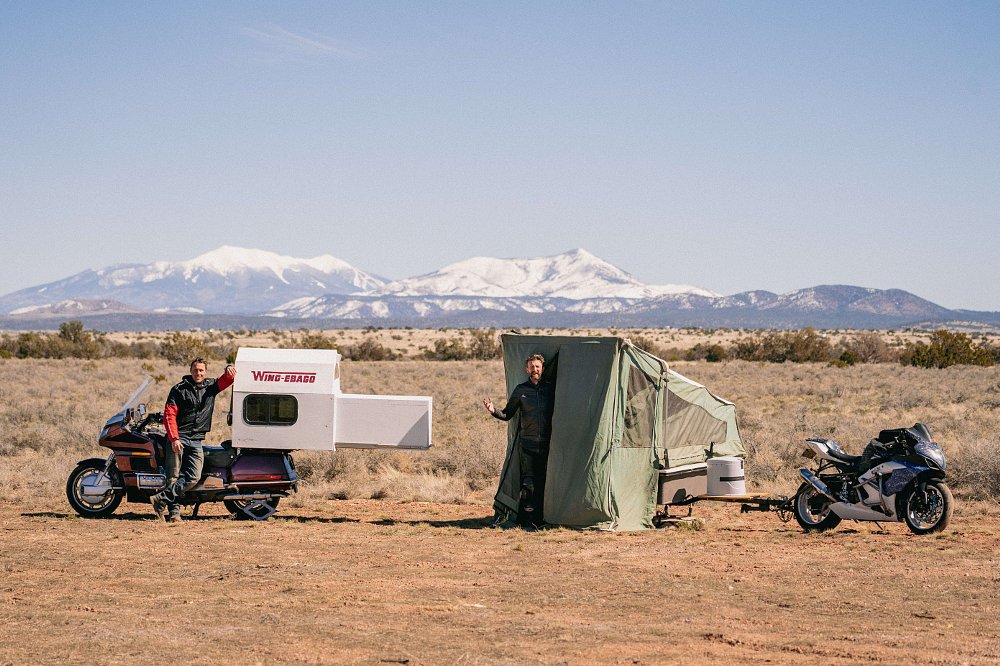 1989 Honda Gold Wing camper and 2006 Suzuki GSX-R1000 towing a trailer, both parked and setup for camping, with mountains in the background.