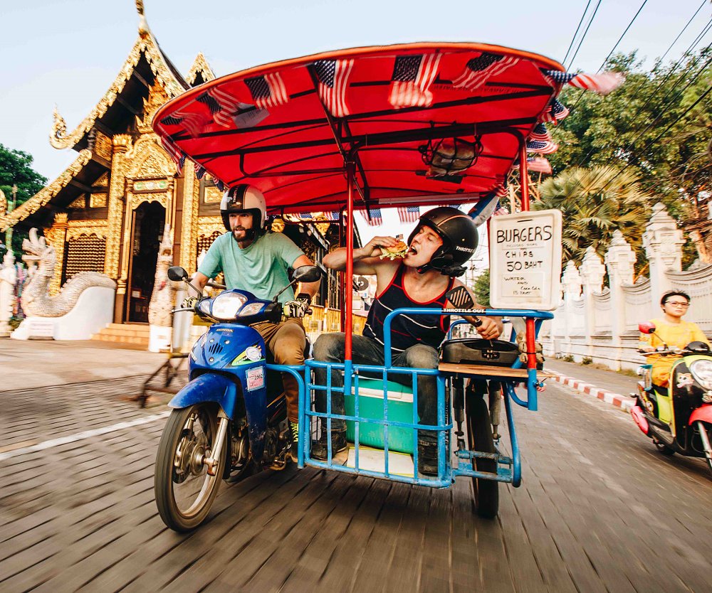 Ari eating a hamburger in a sidecar as Zack drives the scooter