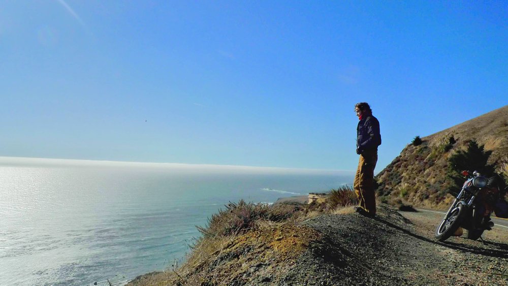 rider overlooking the Pacific ocean on a coastal road with motorcycle parked nearby