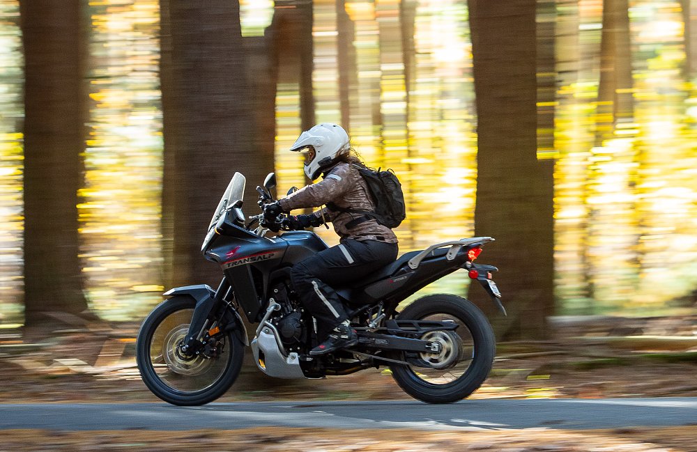 woman riding a Honda Transalp on a dirt road in a sunlit forest