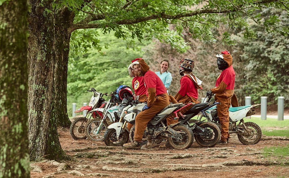 several riders in a wooded area sitting on small motorcycles and smiling, some in costumes
