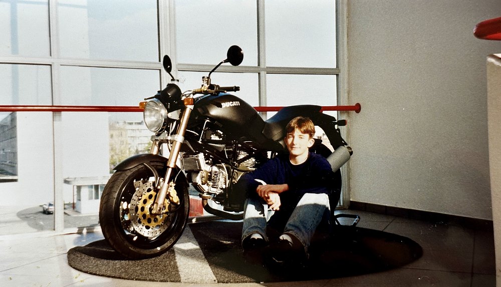 A young man sits next to a black Ducati Monster in the entry of the Ducati Factory Museum, circa 1998.