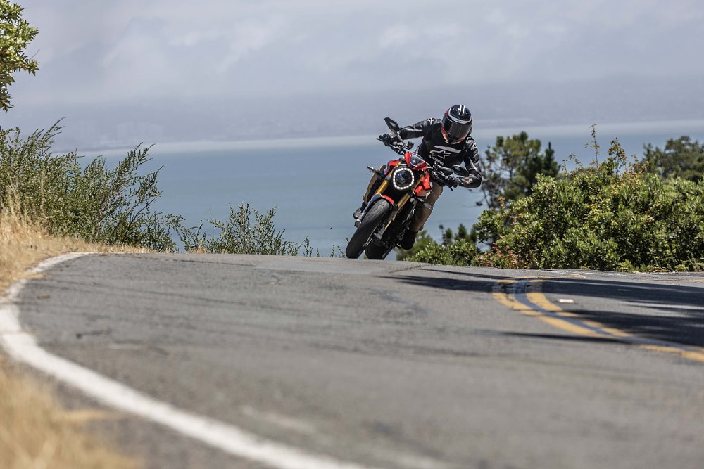 A 2024 Ducati Monster SP rides toward camera on a twisty road, with the San Francisco Bay in the background.