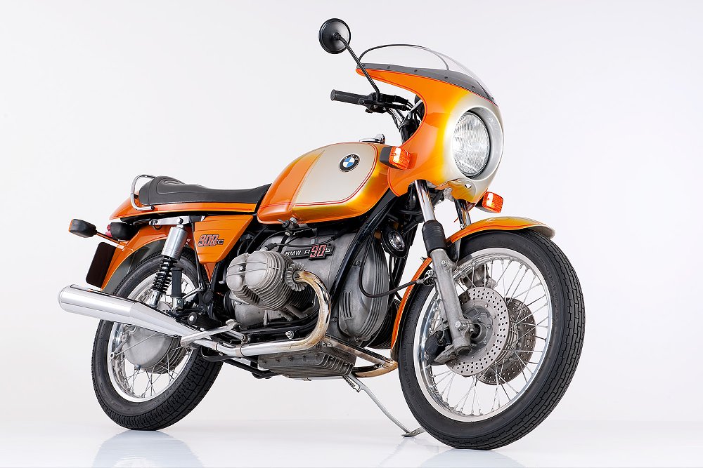 A three-quarters view of a 1975 BMW R 90 S Daytona Orange with a white background.