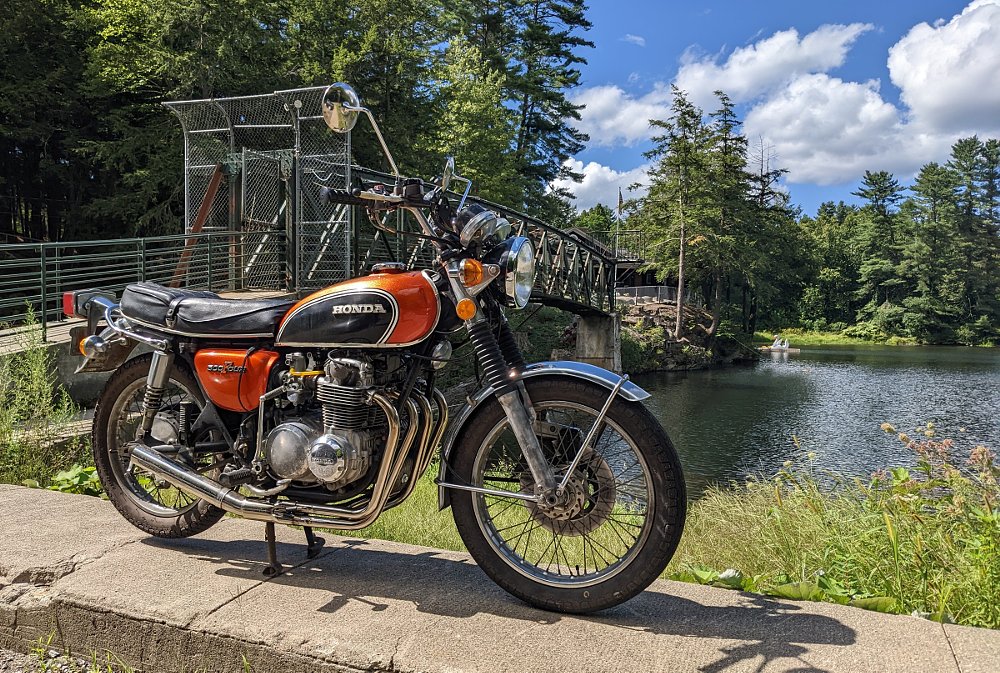 Orange and black 1973 Honda CB500 four-cylinder parked by a forest lake in the countryside