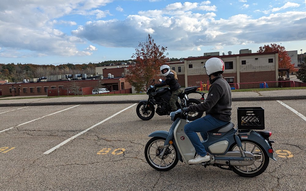 Kate on her Yamaha MT-03 and Joey on the blue Super Cub riding side by side in a parking lot, training