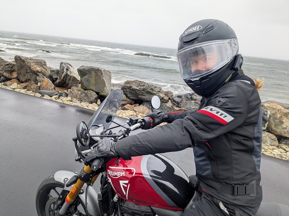 photo of Abby on her motorcycle by the ocean, rain on her helmet, smiling