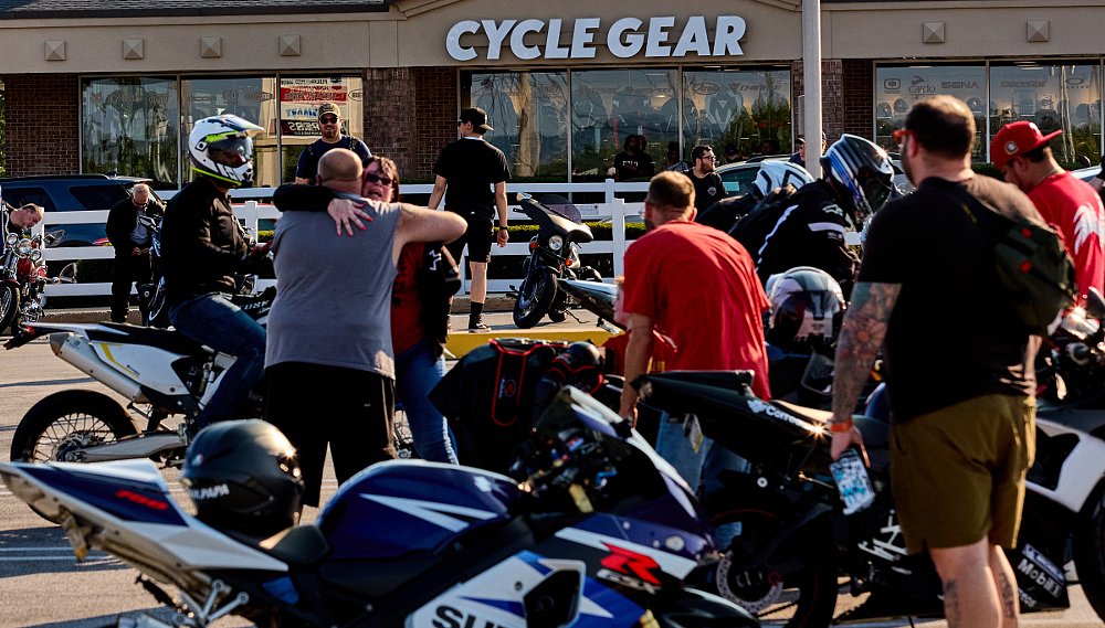 two riders hugging with other riders nearby and many motorcycles parked in front of a Cycle Gear store