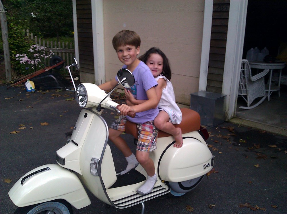 a young Leo sitting on a white scooter with a young girl on the seat behind him