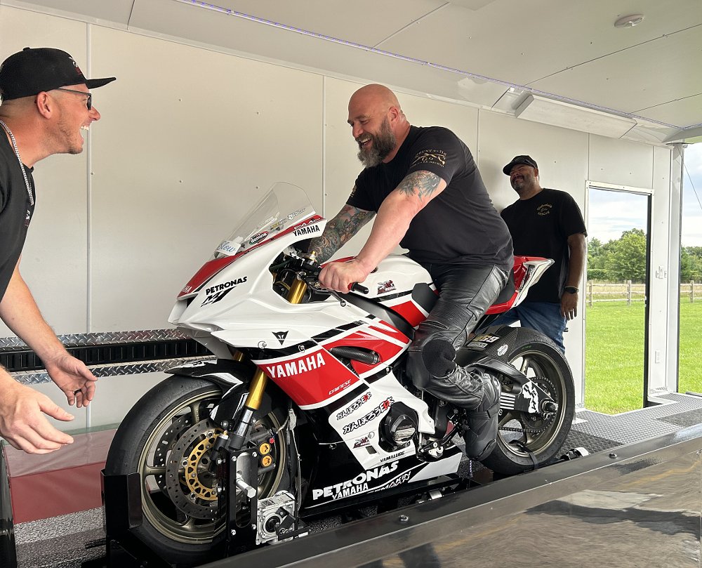 Chadwick, a large man with a beard and tattoos, laughing as he sits on a Yamaha sport bike on a stand