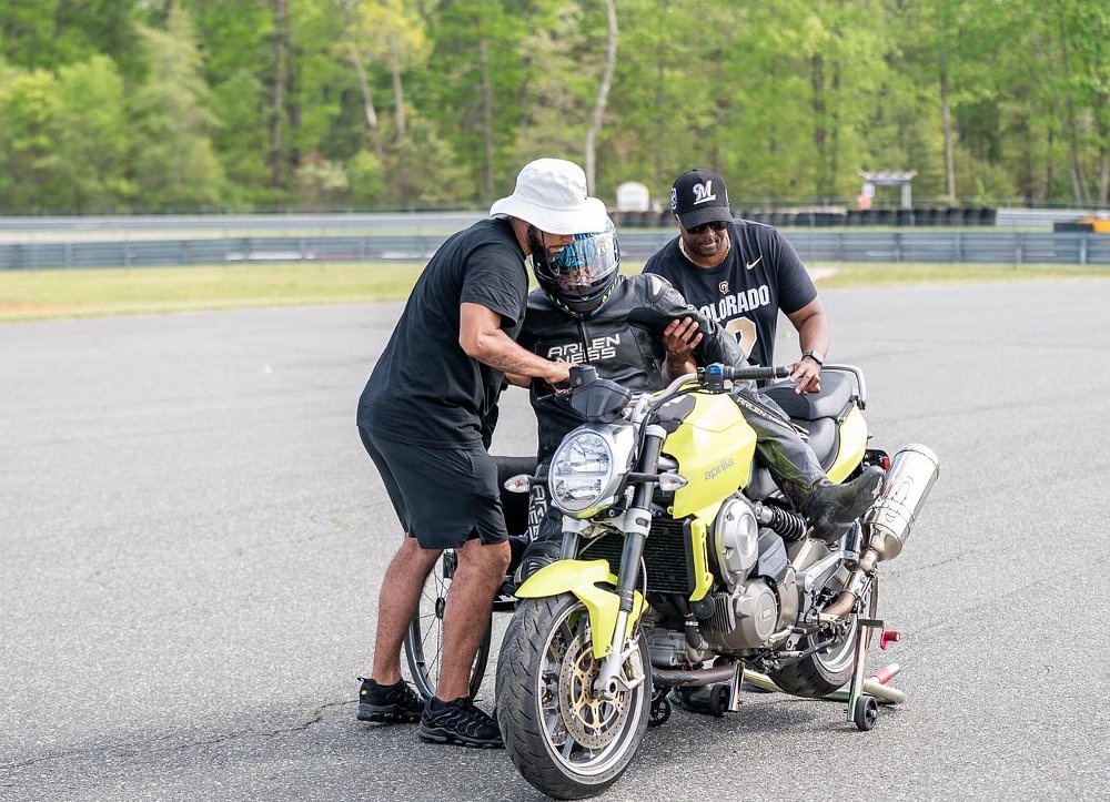 two volunteers help lift a rider onto a yellow motorcycle on a track stand at the track