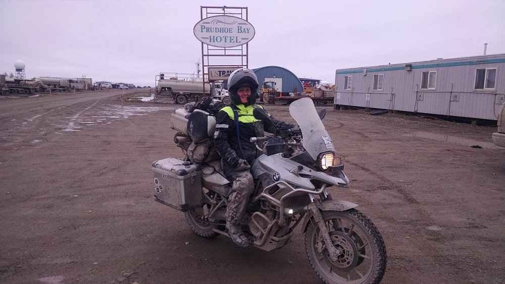 smiling Kate Johnston on her BMW, covered in mud and under a gray sky, in front of the sign at Prudhoe Bay, Alaska