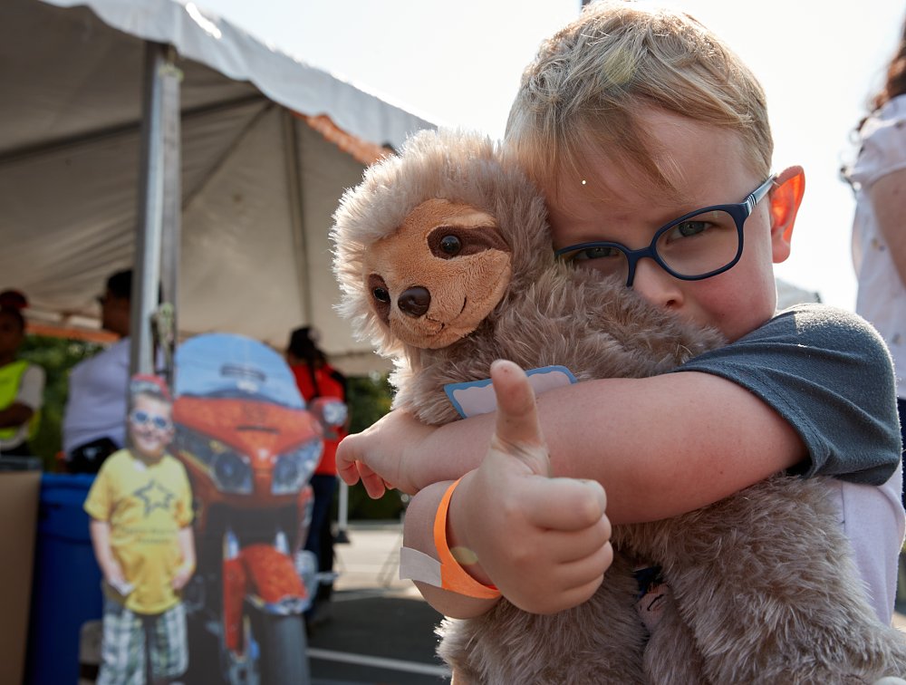 boy giving thumbs up and hugging a teddy bear with a cutout of a motorcycle in the background at a Ride for Kids event