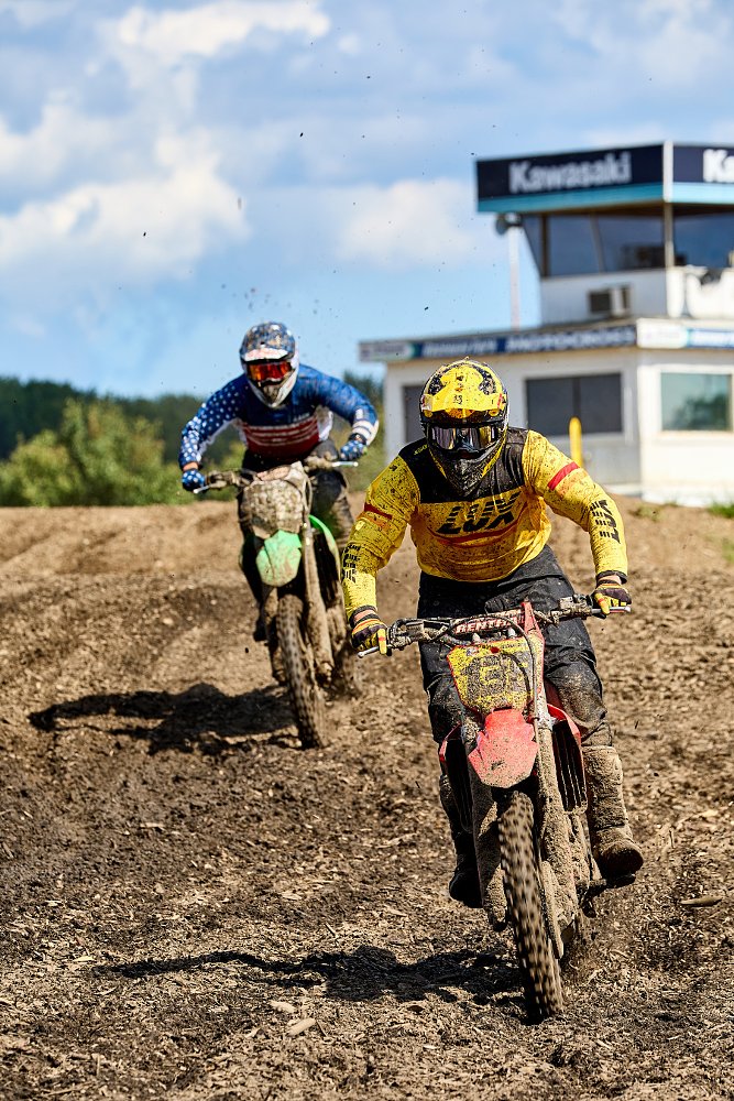 two riders going over jumps on a motocross track with a small scoring tower in the background