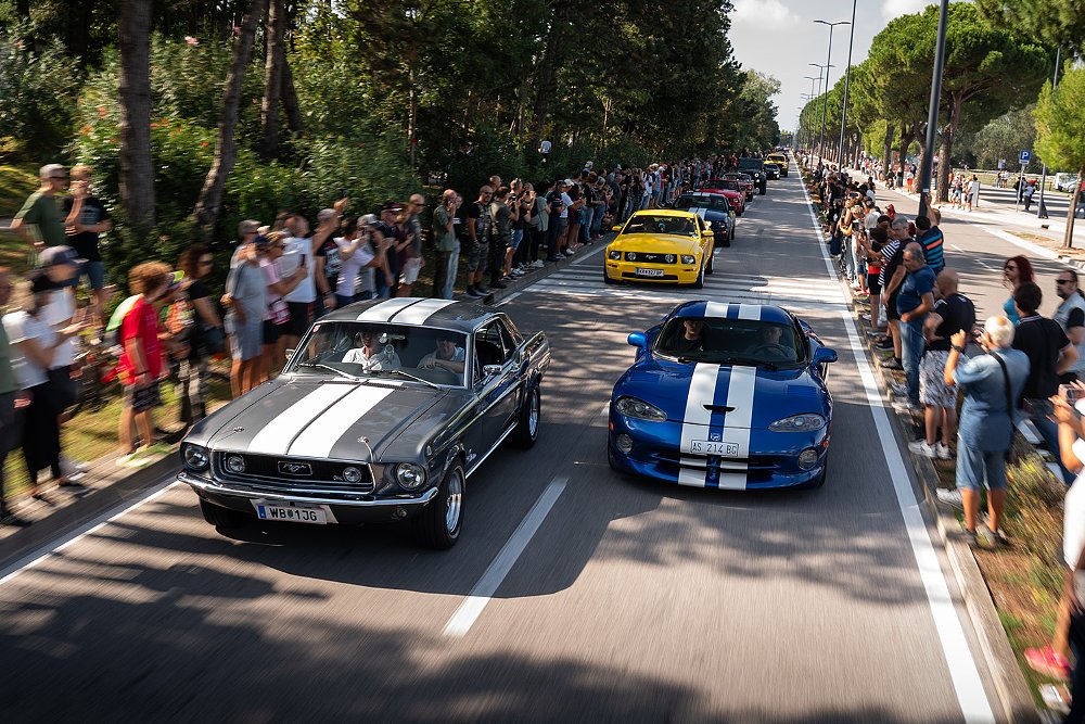 American muscle cars and classic cars drive by rows of onlookers.